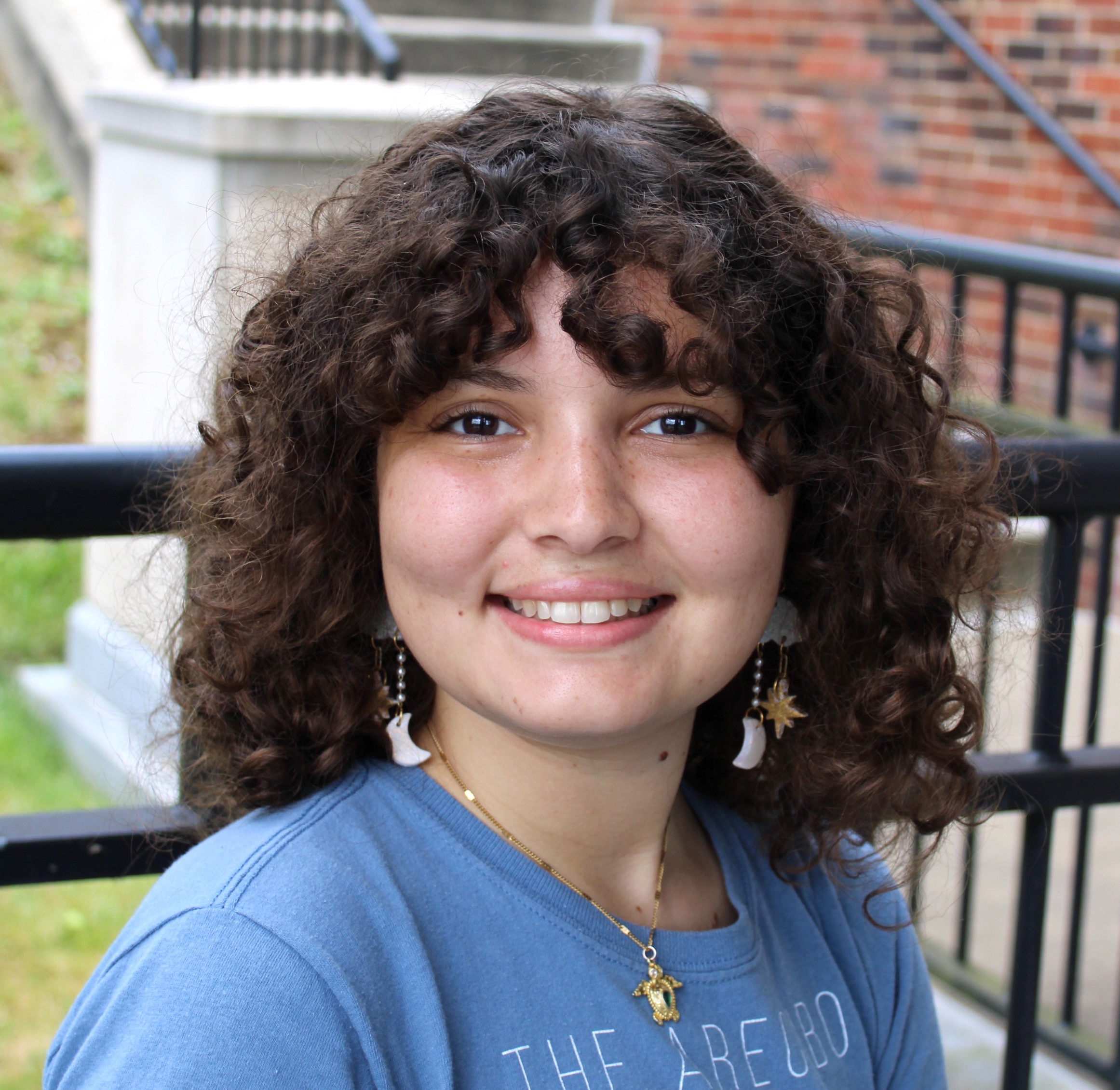 Smiling young woman with chin length curly brown hair and dark brown eyes wearing moon and star earrings and a blue tshirt.